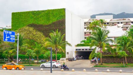 Sanya, Hainan island, China - May 15, 2019: Road traffic. Chinese yellow taxi on the street of Sanya. City landscape. A building with an unusual green facade from living plantsのeditorial素材