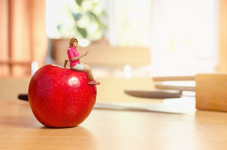 a petite woman reading a book sitting on a red apple in the kitchen on a wooden tableの写真素材