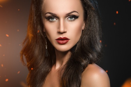 woman standing in the sparks of fire. Beauty portrait of brunette in black earrings on a dark background with the flying sparks from the fireの写真素材