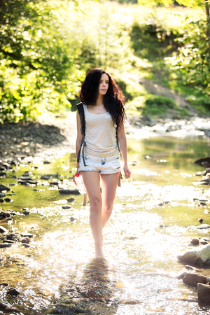 Outdoor shot of attractive young woman with backpack standing in a mountain stream. Female hiker in creek water.Portrait of young pretty brunette woman holding bottle of water.の写真素材