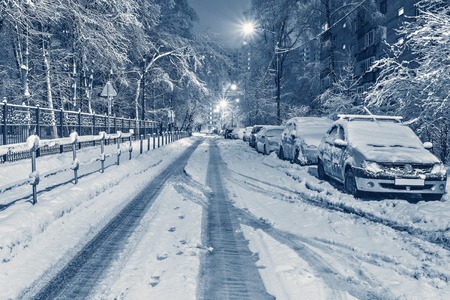 Cars under the snow on the night winter city street.