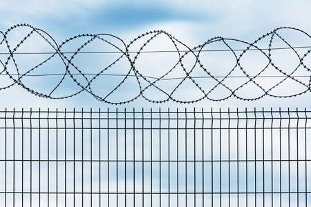 Metal fence with barbed wire on gray sky background.の写真素材