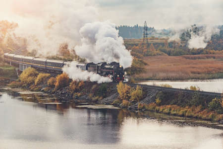 Retro steam train moves along the lake at autumn sunrise.