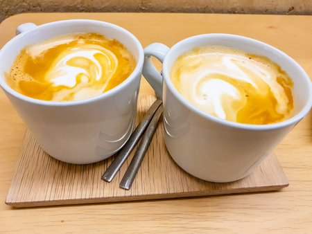 Two cups of latte with its foam drawing shapes. Presented on a wooden board in precious warm tones.の写真素材