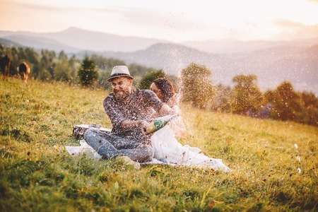 loving couple sitting on a plaid in a field on a background of mountains. opened champagne sprayed.の写真素材
