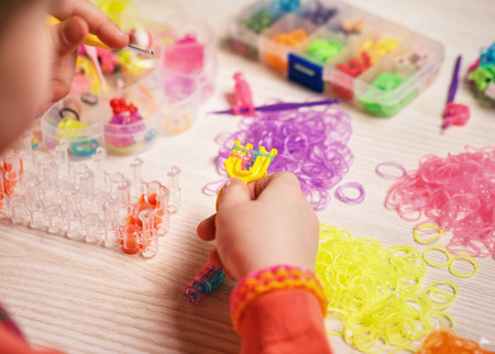 extracurricular activities, group, education and handwork concept - hands weaving of colored rubber band bracelet, closeup,wooden backgroundの写真素材
