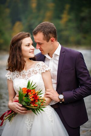 The newlyweds look into each other's eyes, standing on the river bank. A bride with a with a bouquet of live flowersの写真素材