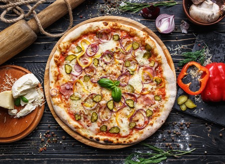 Original pizza with onion, pickled cucumber and ham. Concept promotional flyer. Pizza on a black wooden background among spicesの写真素材