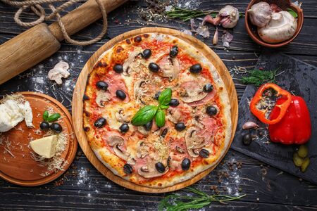 Exquisite pizza with ham, mushrooms and olives. Poster for Restaurants or pizzerias. Pizza on a black wooden background among spicesの写真素材