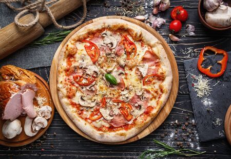 Pizza with ham, mushrooms, tomatoes and leaf of basil. Pizza is on the table among the ingredients for its preparation. Pizza on a black wooden background among spicesの写真素材