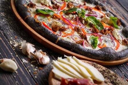 Italian pizza with black dough with cheese, pepper, onion and basil on a wooden tray. Ingredients of pizza on the foreground.の写真素材