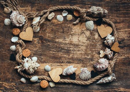 Frame made of rope, various seashells and wooden hearts on old burnt wooden background. Place for text. Sea travel concept. Top view.の写真素材