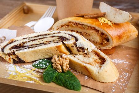 Poppy and apple strudel on a rustic wooden tray. The scent rolls decor consists of cinnamon, mint, nut, lemon peel and powdered sugar. Close up, blurred background.の写真素材