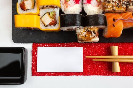 Different types of sushi rolls with a variety of stuffing on a dark stone board and soy sauce on a white table. Blank white card for text or inscription. Promotional shooting. Top view.の写真素材