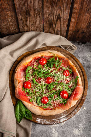 Italian pizza with a crispy crust on a round wooden tray. Pizza with prosciutto arugula and cherry tomatoes. Top view.の写真素材