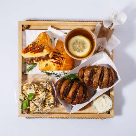 Lemon tea, salad, baked potatoes with meat, sauce and rosemary sausage rolls. Dishes are served with disposable utensils on a rustic wooden tray. Delicious lunch. Top view. Street food concept.の写真素材