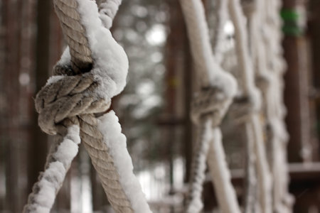 A close-up view of a rope knot covered in snow, with elements of a rope park in the blurred...