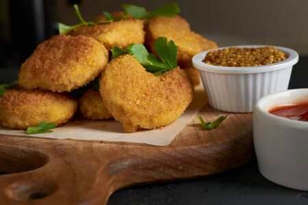 Appetizing chicken nuggets on a cutting board with parchment paper sprinkled with parsley leaves and tomato sauce and mustard seeds.の写真素材