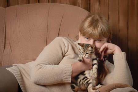 Cute middle aged Caucasian woman in knitted sweater lying on sofa looking at camera holding tabby kitten indoorsの写真素材