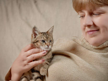 Pretty woman in a beige sweater holds a tabby kitten in her hand indoors. Close up from low angle...