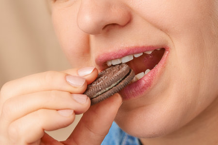 Close up happy Caucasian woman holding chocolate cookie in her hands and biting it. Three-quarter...