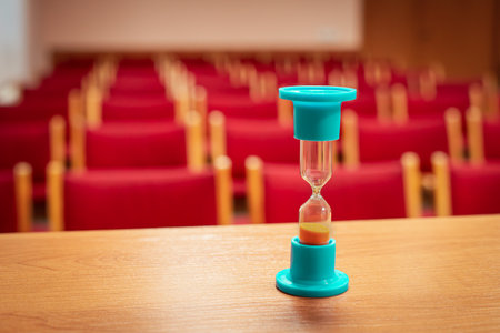 Hourglass on a table in an auditorium with chairs. Indoors.