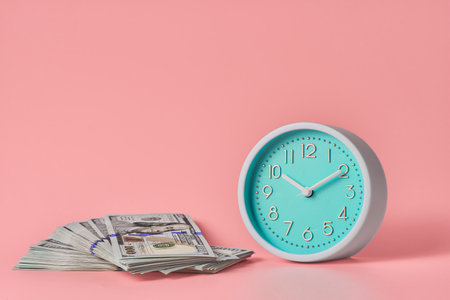 Round analog clock and a stack of many hundred dollar bills on a pink background with copy space.の写真素材