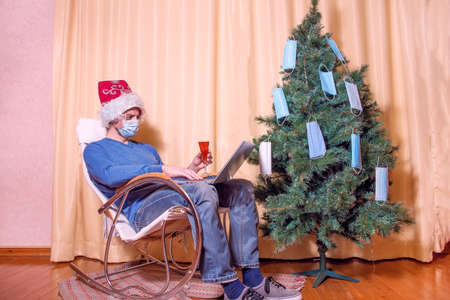A man in a Santa Claus hat and a medical mask sits near a Christmas tree. A man aged 40 is...