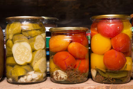 Jars of canned vegetables are on the shelf. Pickled cucumbers and tomatoes.