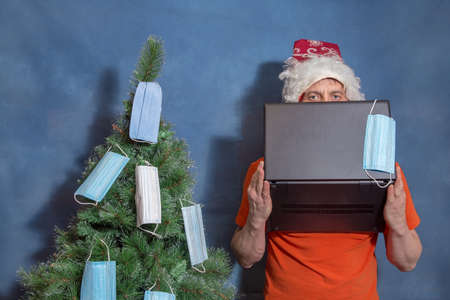 A man wearing a Santa Claus hat covers his face with a computer. Medical masks hang on the Christmas...