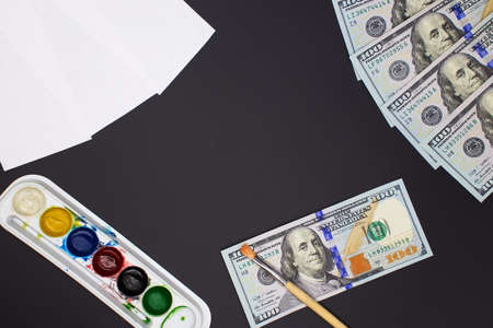 Dollar bills, paints, brushes, paper lie on a black background.