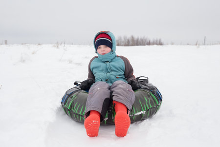 A boy, 5 years old, sits on a cheesecake for downhill skiing. Open air games.