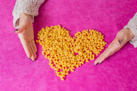 Pink background. A heart is made of pasta. Women's hands.の写真素材
