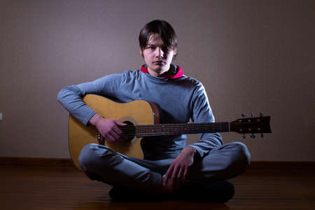 A young man is sitting on the floor and holding a guitar ..