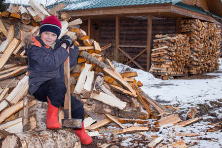The boy sits next to a pile of firewood and holds a large ax in his hands.の写真素材