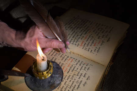 A man's hand holds a pen for writing. Burning candle. Old book with open pages. The concept of mysticism and secret knowledge, esotericism. Selective focus on the hand.の写真素材