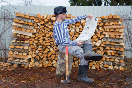 A country man sits on a tree stump and reads a newspaper. There is an ax nearby. In the background...