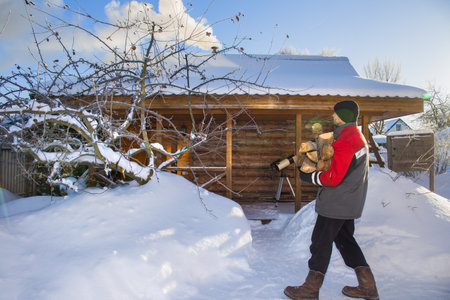 A European man carries an armful of firewood to the bathhouse. In the background is a village bathhouse. Smoke is coming from the chimney.の写真素材