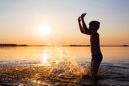 Photo of a person in motion. Silhouette of a man against the background of the sun. A child splashes water in the lake.の写真素材