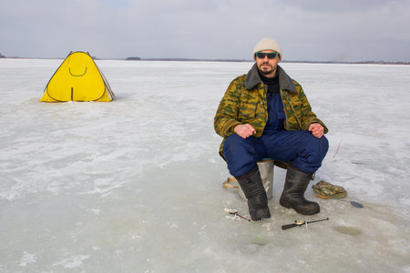 A man fishes on the ice of a lake. A fishing tent stands on the ice of a lake.