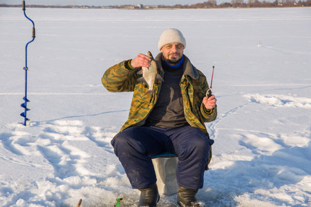 A European man, 49 years old, is fishing on the ice of a frozen lake. holding caught fish in his hands. looks at the camera.の写真素材