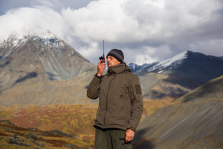 A man, European, 50 years old, stands with a walkie-talkie in his hands against a backdrop of mountain peaks. He's talking on the radio.の写真素材