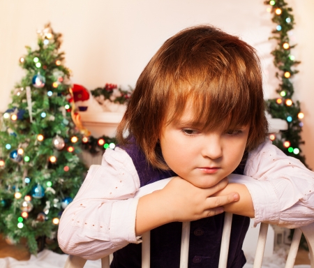 Sad kid sitting with Christmas tree and fire place on the background sad as Christmas is overの写真素材