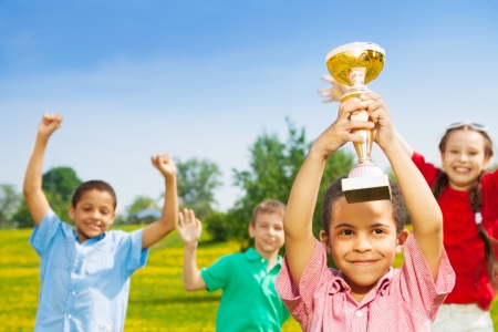 Close shoot of black happy smiling little boy holding prize cup with group of boys and girls on backgroundの写真素材