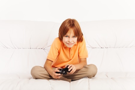 Happy Caucasian boy playing video games holding game controller sitting on the coach in living room