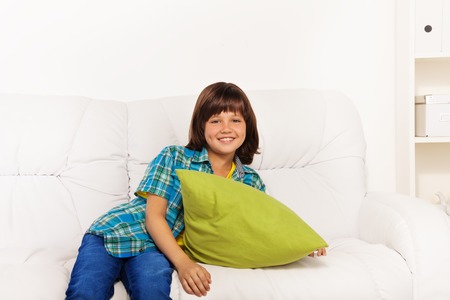 One happy calm and relaxed little boy 6 years old sitting with green pillow on the white leather coach in living room at homeの写真素材