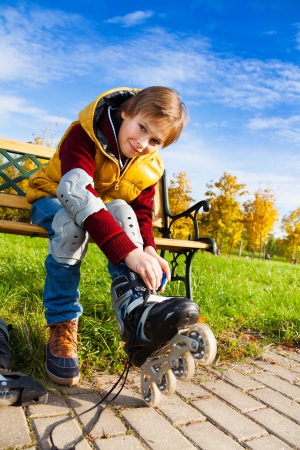 Close portrait of 10 years old boy in casual autumn clothes putting on roller skates sitting on the bench in the parkの写真素材
