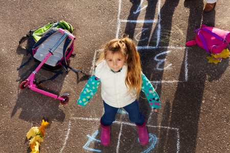Nice little girl jumping over hopscotch game after school with bags and scooter laying nearの写真素材