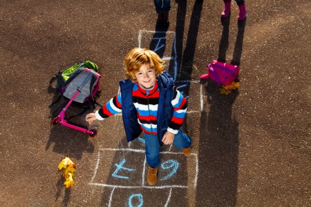 Nice blond boy jumping over hopscotch game after school with bags and scooter laying nearの写真素材