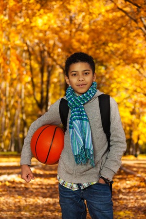 Handsome black boy 10 years old standing in the autumn park under maple trees with orange basketball...